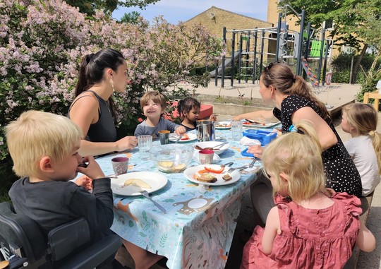 børn og voksne spiser frokost udenfor ved et bord på børnehusets legeplads