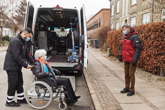 Kvinde i kørestol hjælpes ind i flextrafikbil mens en pårørende mand ser til